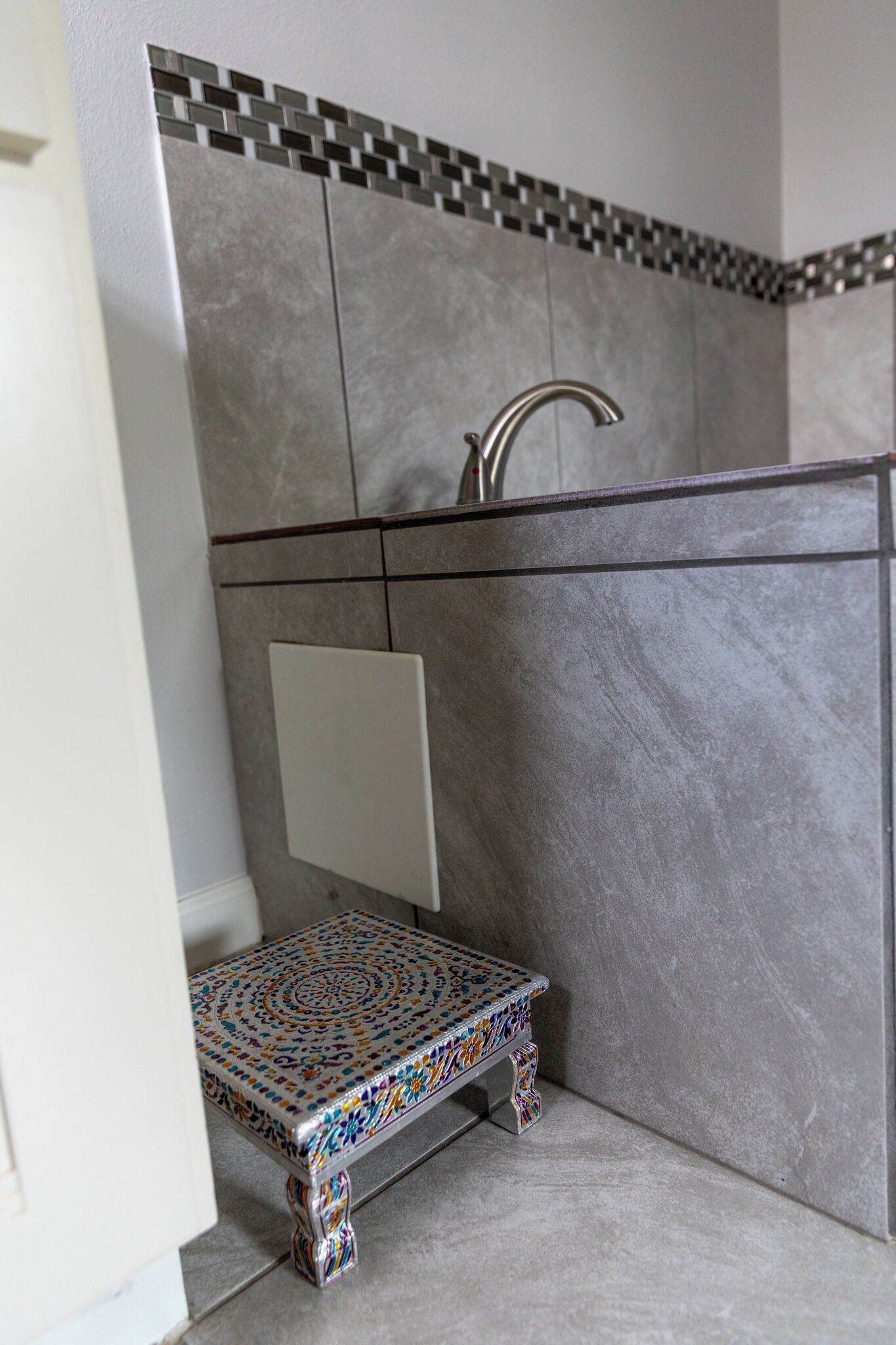 Et badeværelse med en lille, farverig bænk, en moderne vandhane og et flisebeklædt backsplash.