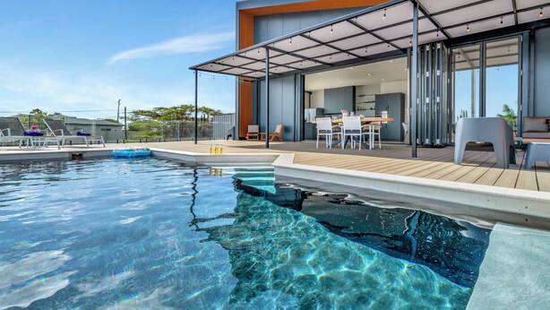 Un área de piscina exterior moderna con muelle de madera, sillas de descanso y una zona de estar cubierta con mesas y sillas.
