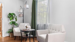 A room with two white armchairs, a round table with a decorative item, a floor lamp, a large green plant, and a window with sheer curtains. - Newly active beautiful 5-bedroom house in a prime location near DC (Arlington)