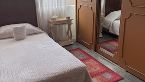 A neatly made bed with a white quilt, a bedside table with a tissue box, a small wooden nightstand, and a red and white patterned rug.