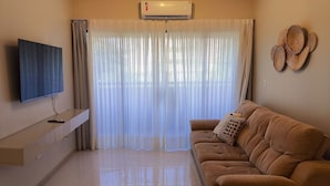A living room with a flat-screen TV, a sofa, a TV stand, and a large window with sheer curtains. - Tarantus (Joao Pessoa)