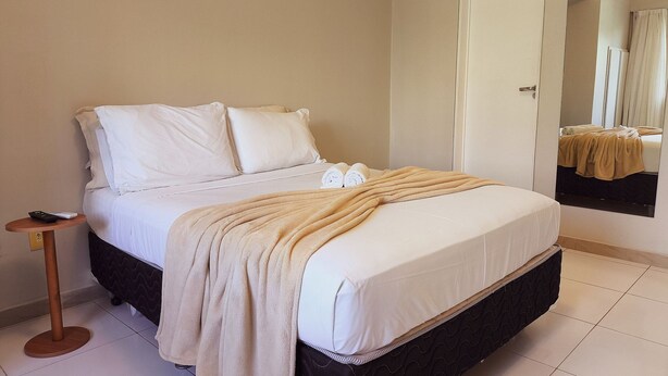 A neatly made bed with white linens and a beige blanket, a bedside table with a remote control, and a mirror reflecting the bed and a bathroom. - Tarantus (Joao Pessoa)