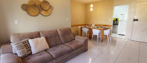 Una sala de estar con un sofá marrón, una mesa de comedor con sillas, una cocina al fondo y una puerta.