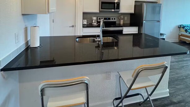 A modern kitchen with white cabinets, a black countertop, stainless steel appliances, and two bar stools. - Pleasant apartment in Austin (Leander)