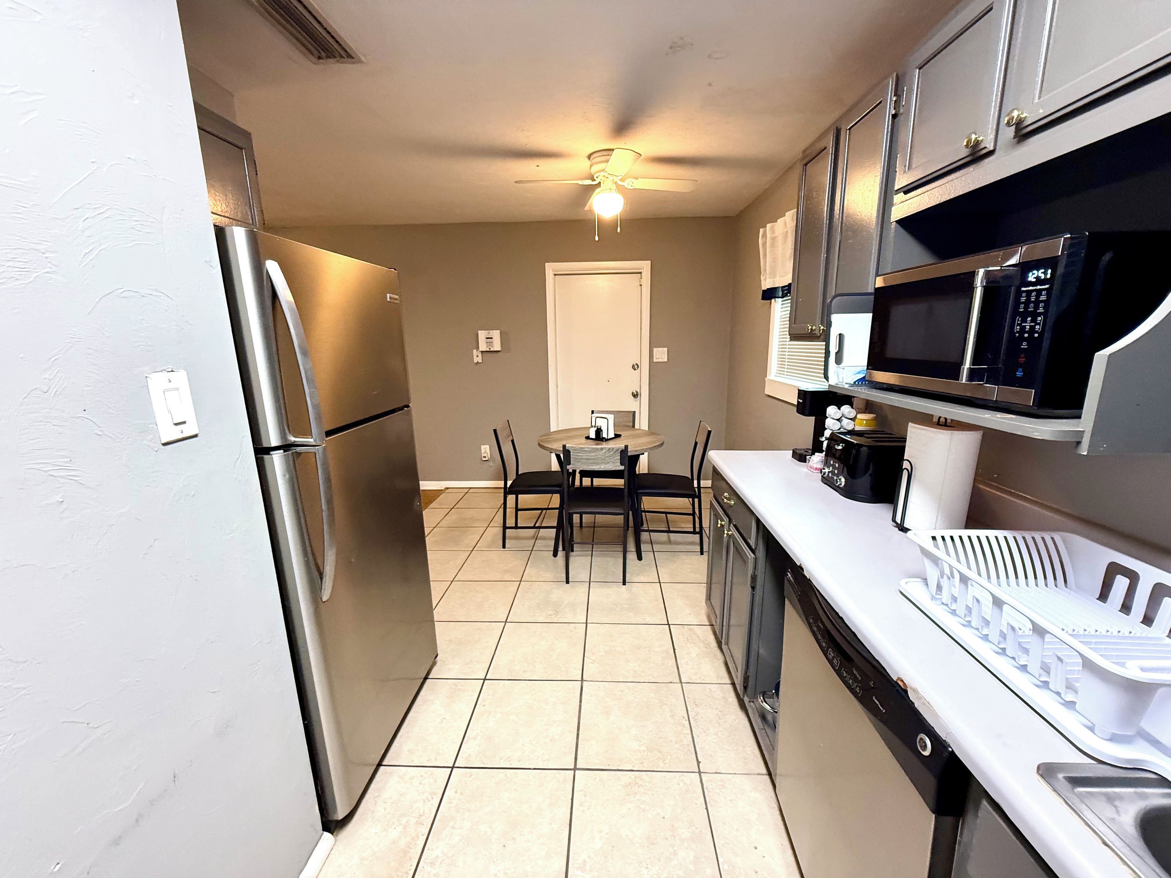 A compact kitchen with a refrigerator, microwave, and a dishwasher. There is a dining table with chairs and a ceiling fan.