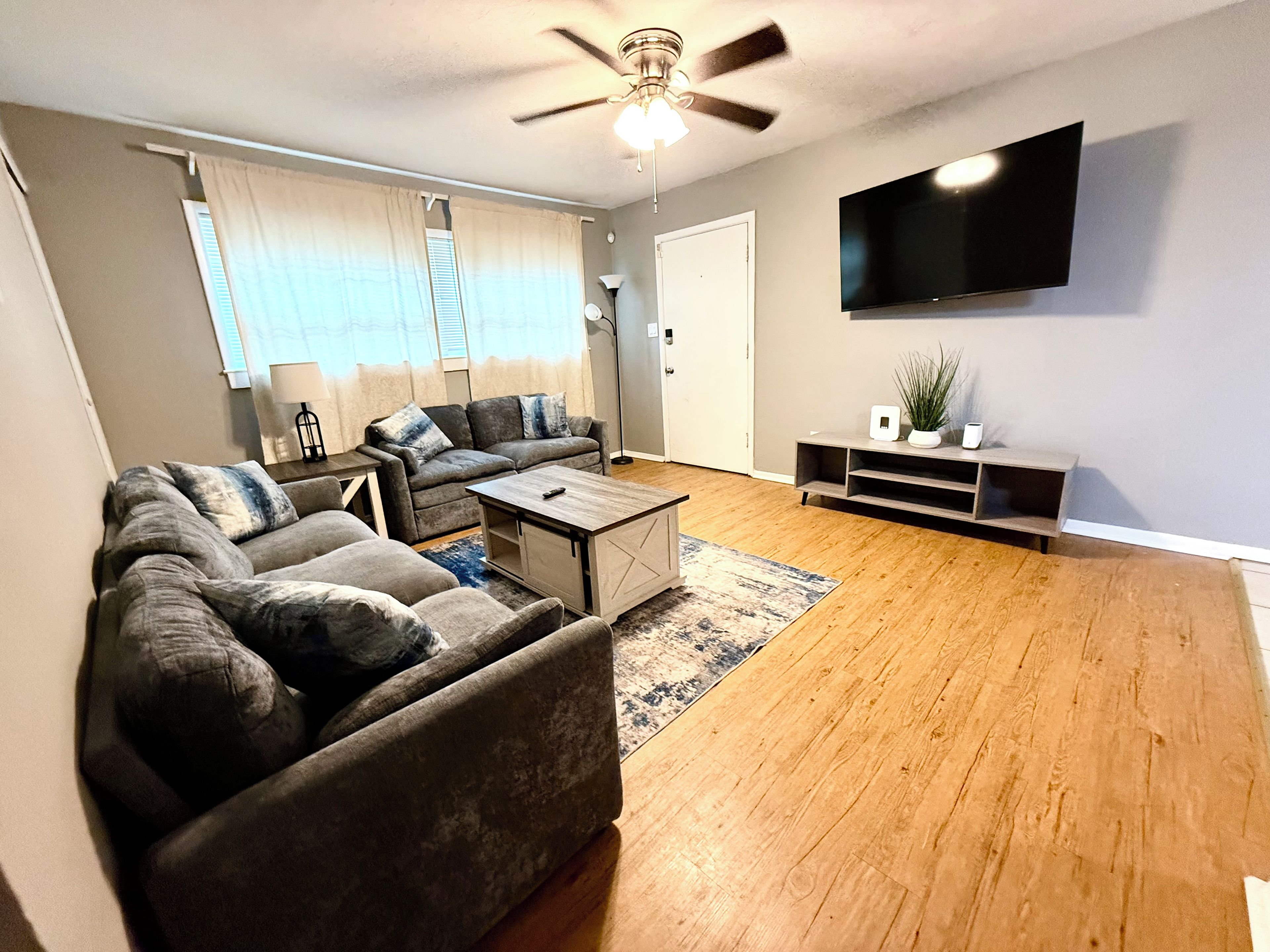 A living room with a sectional sofa, a coffee table, a flat-screen TV mounted on the wall, and a ceiling fan.