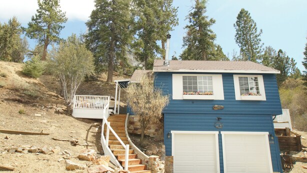 Uma casa térrea azul com uma escadaria de pedra que leva a um deck, cercada por árvores e uma encosta.