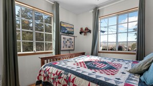 Quarto com uma cama grande, cabeceira de madeira, colcha, duas janelas com cortinas, TV na parede e vista para árvores e montanhas.