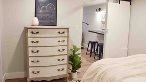 Un dormitorio con una cama, un aparador con tiradores florales, una planta en maceta y un cuadro en la pared.