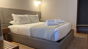 Ein ordentlich bezogenes Bett mit weißen Leinen und einer grauen Tagesdecke, ein Nachttisch mit einer Lampe und eine Holzbank.