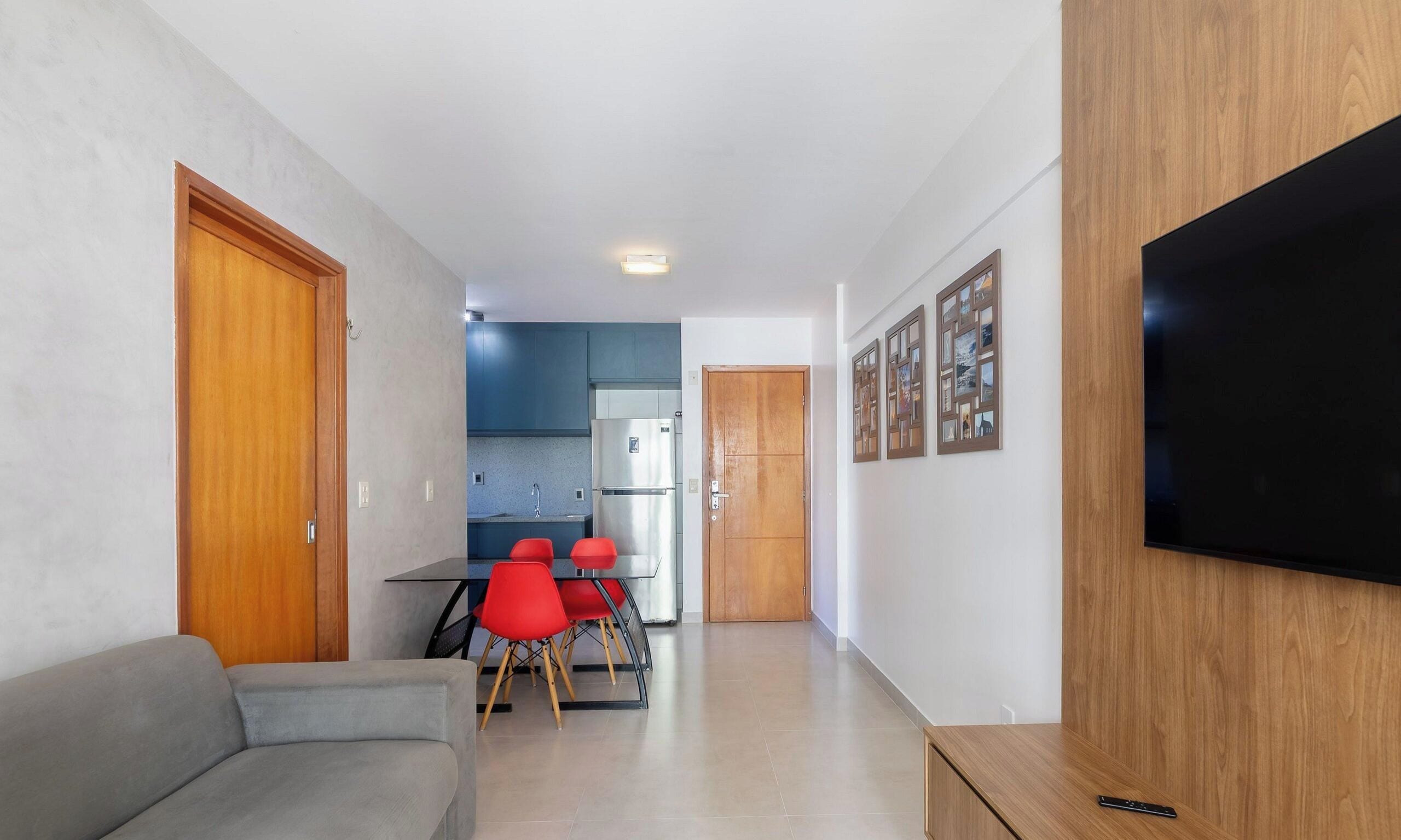Una sala moderna con un sofá, una zona de comedor con sillas rojas, una cocina con armarios azules y un televisor de pantalla plana montado en la pared.