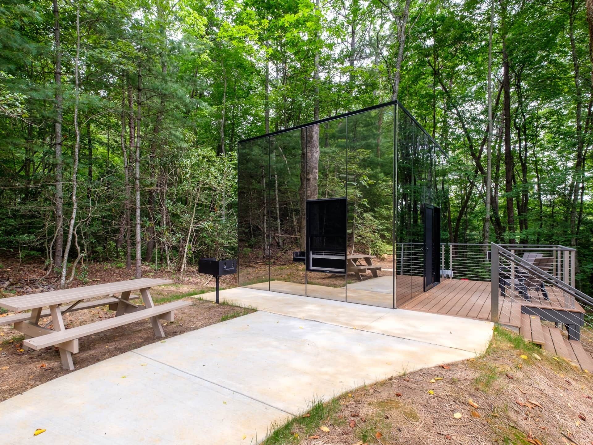 A modern glass structure in a forest setting with a picnic table and a wooden deck.