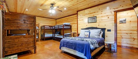 Un dormitorio de cabaña de madera con literas, una cama individual, una mesita pequeña y un ventilador de techo.