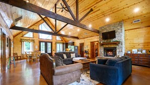 A spacious living room with wooden beams, a stone fireplace, and a television mounted on the wall.