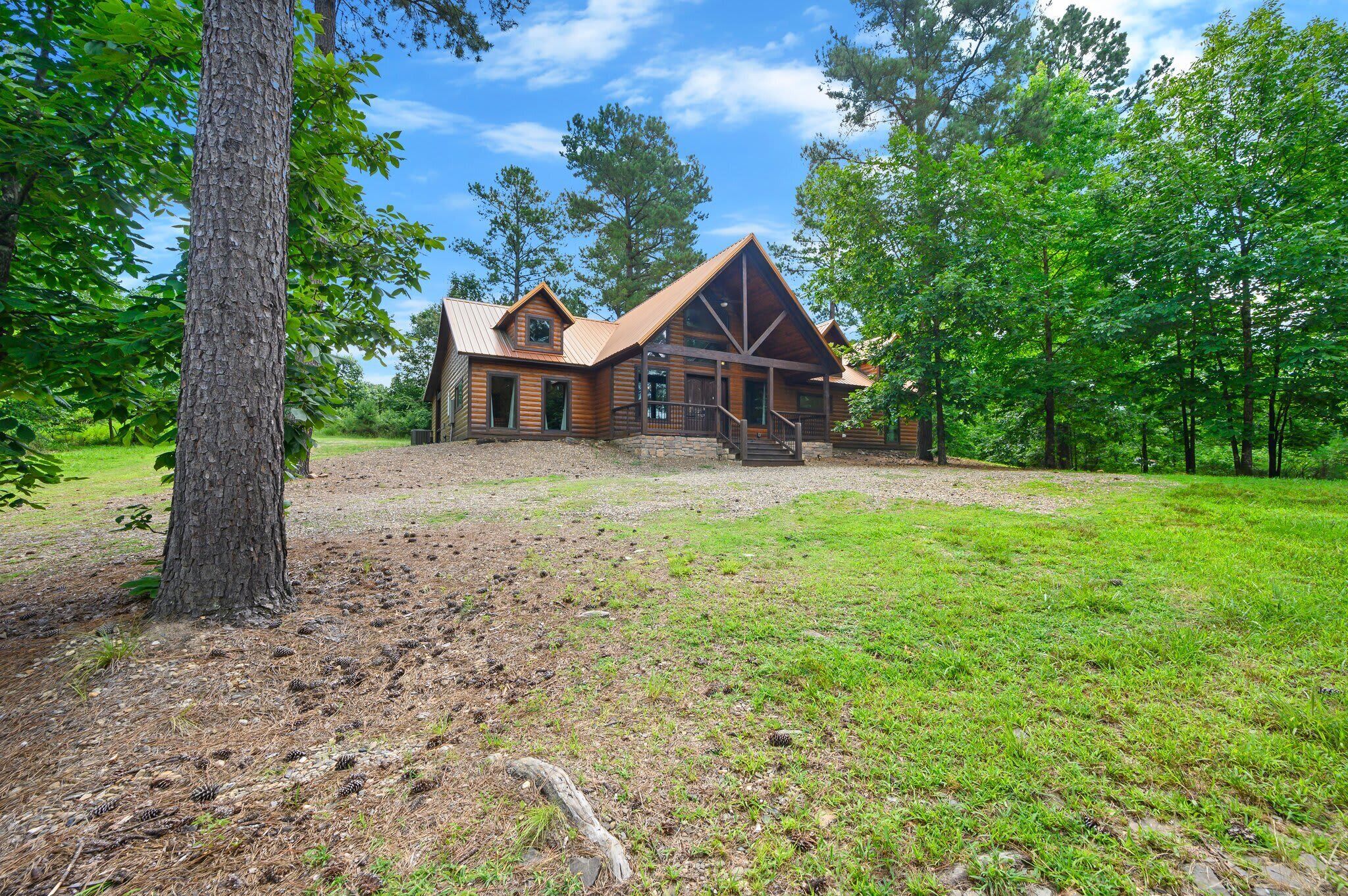 A rustic log cabin with a prominent front porch and a large window, surrounded by a grassy area and trees.