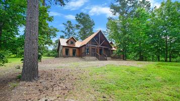 A rustic log cabin with a prominent front porch and a large window, surrounded by a grassy area and trees.