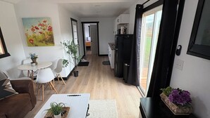 A spacious living area with a white coffee table, a brown sofa, and a dining table with chairs. There is a kitchen area with a refrigerator and a window with curtains. A painting is visible on the wall.