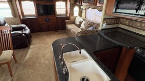 A compact RV kitchen with a sink, stove, and microwave. There is a seating area with a couch and chairs. - Cool RV with AC in Bayfield next to small lake (Bayfield)