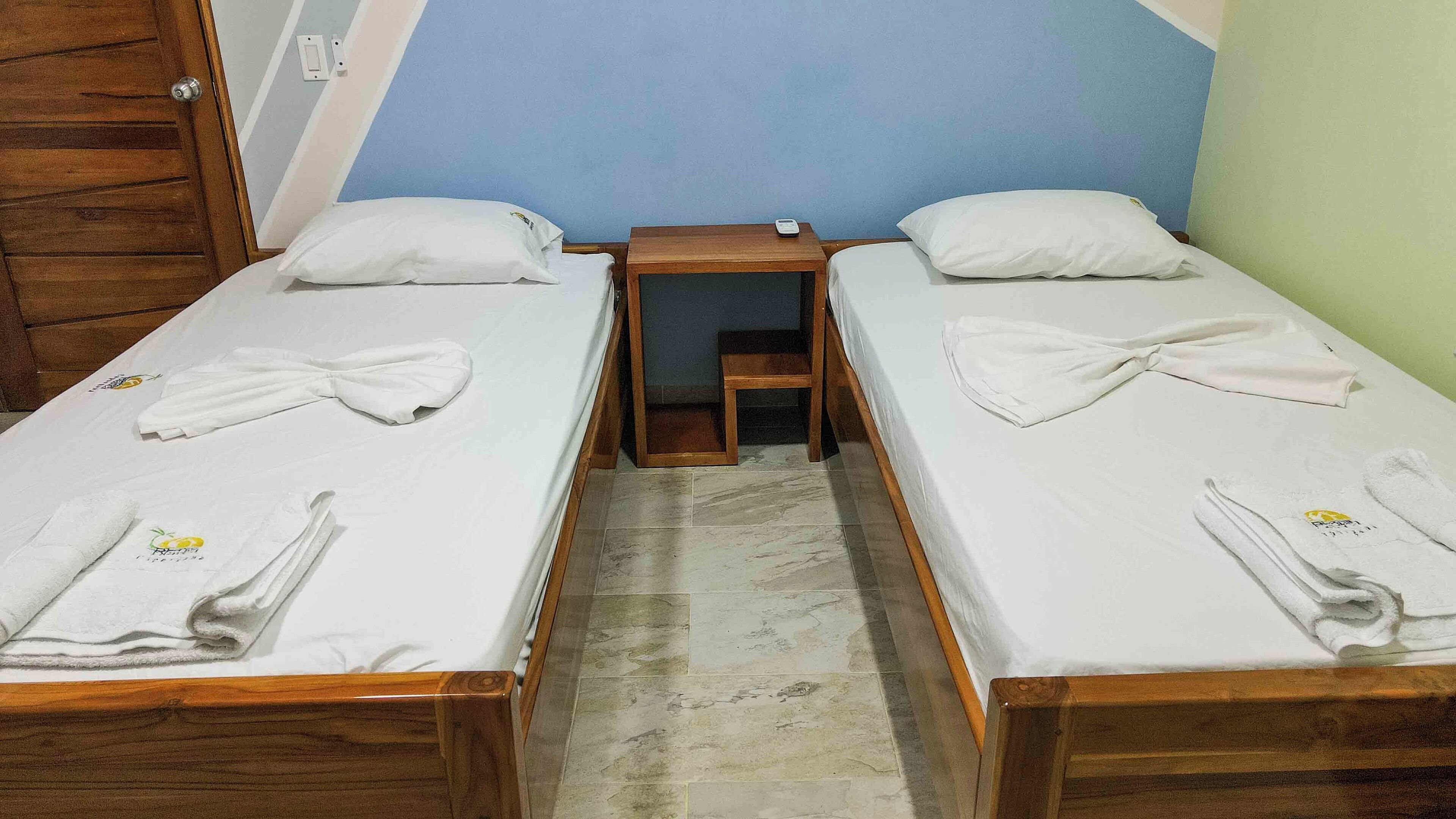 Une chambre avec deux lits simples, chacun doté d’un cadre en bois, de draps blancs et de serviettes soigneusement pliées. Une petite table de chevet en bois se trouve entre les deux lits.