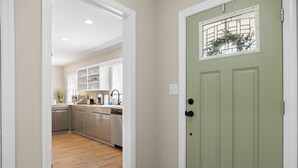 Un corridoio conduce a una cucina con pavimento in legno, mobili bianchi e una porta verde con finestra.