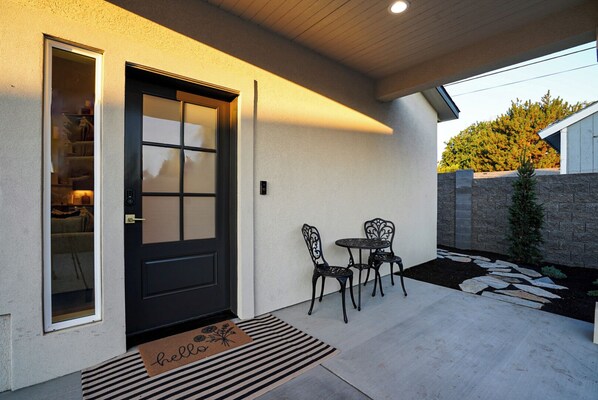 A modern house exterior with a black door, a small patio with two chairs, and a stone pathway.