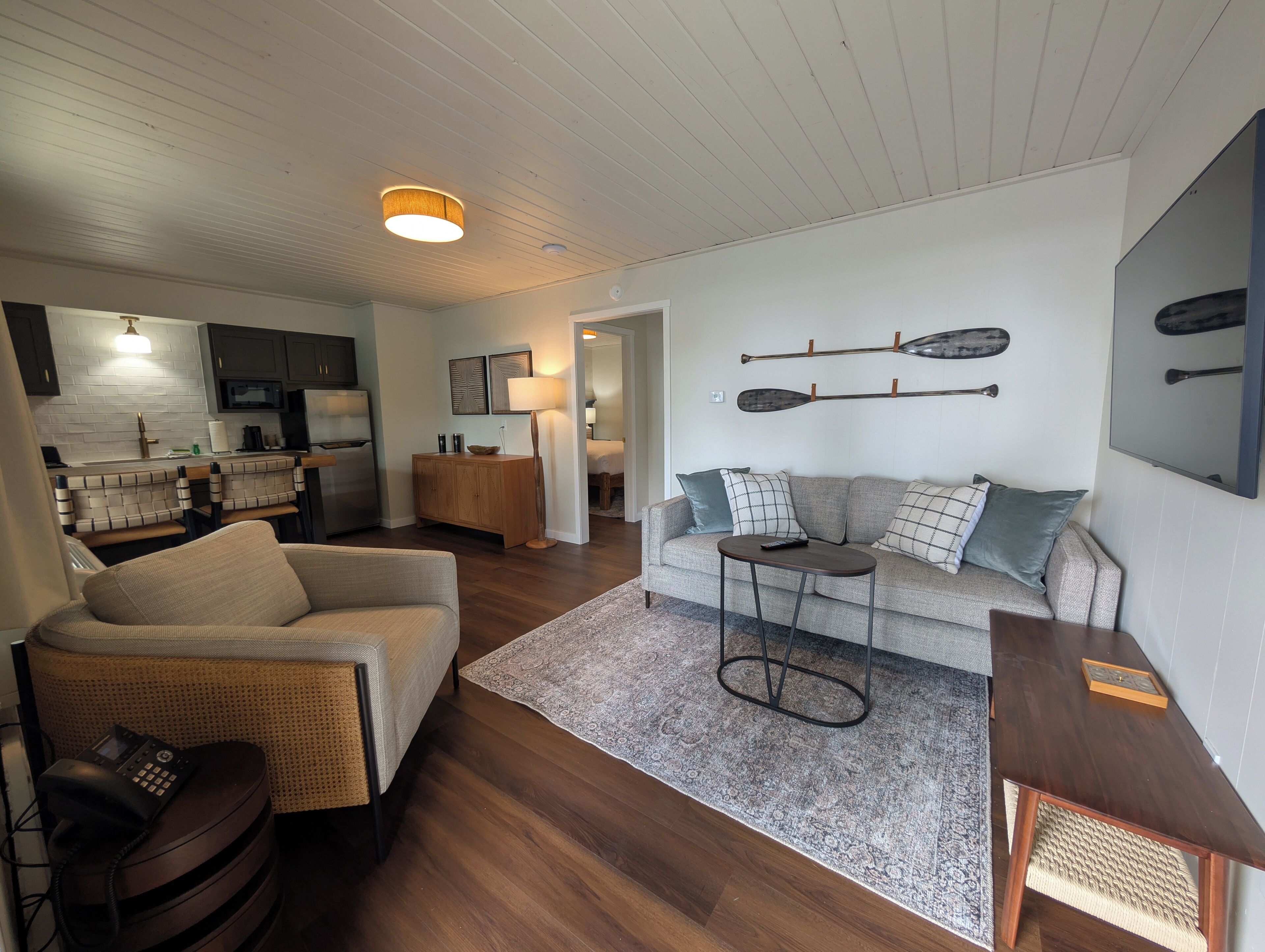 A living room with a sofa, a coffee table, and a wicker chair. There is a kitchen area with a refrigerator and a microwave.