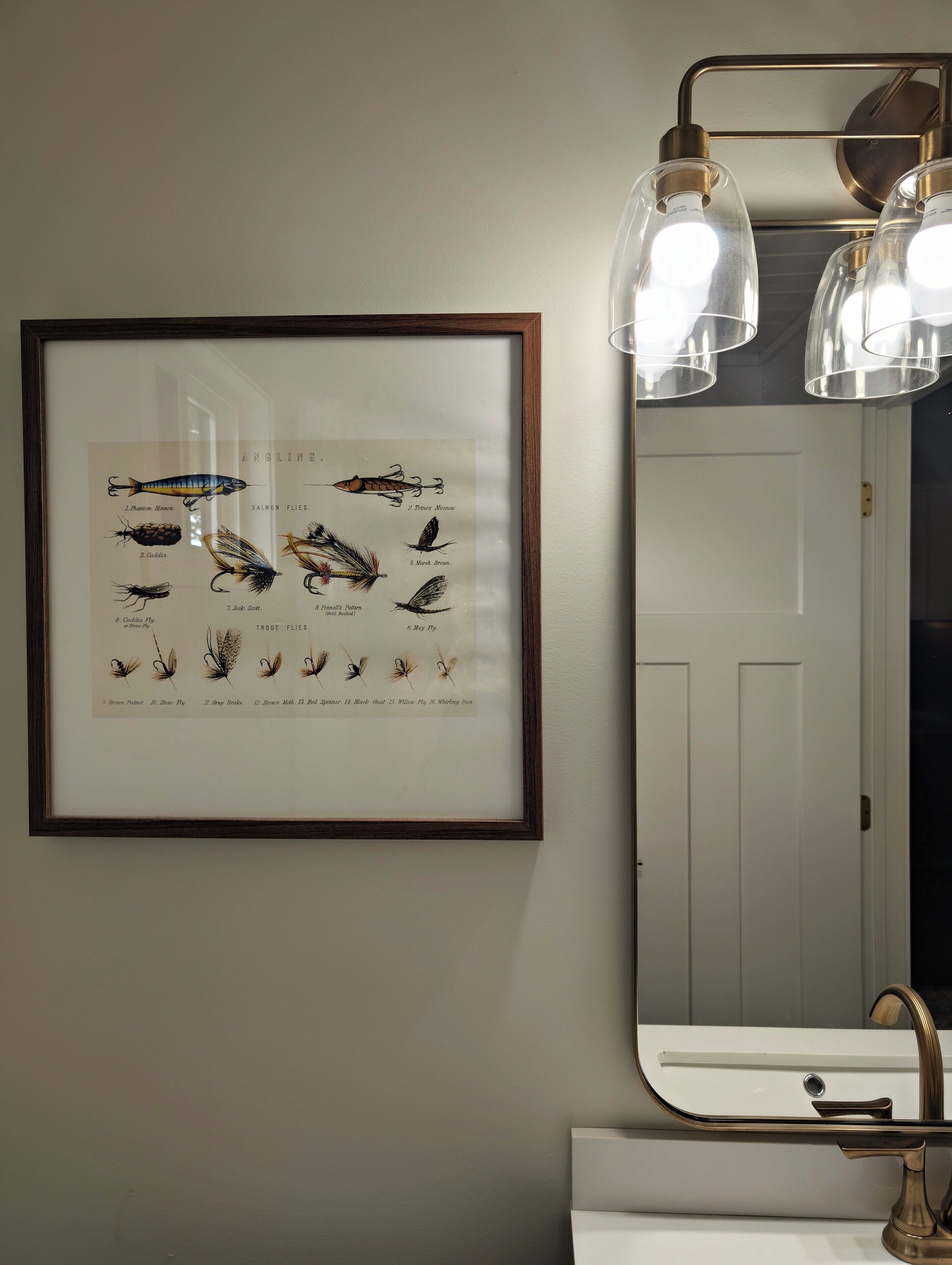 A framed angling flies chart on a wall, a mirror with a gold faucet, and a white sink.