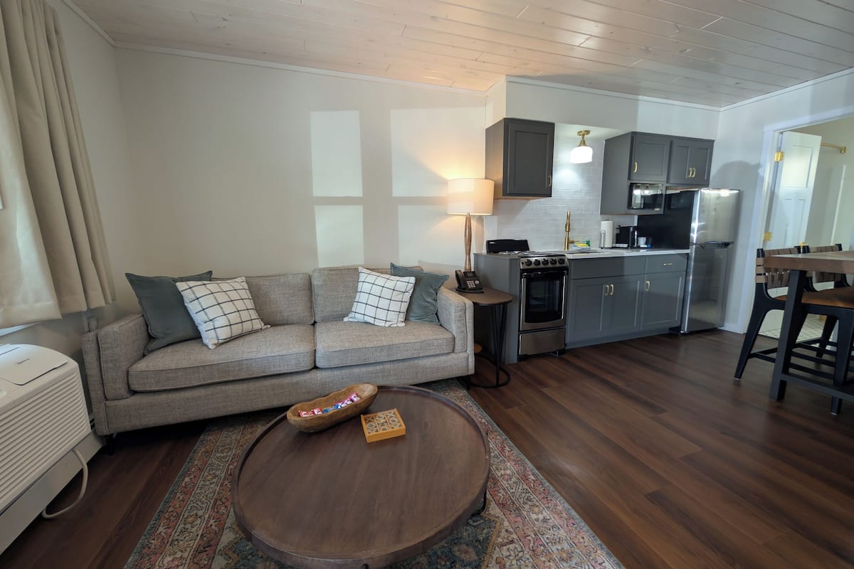 A modern living room with a sofa, coffee table, and a small kitchen area with a stove and refrigerator.