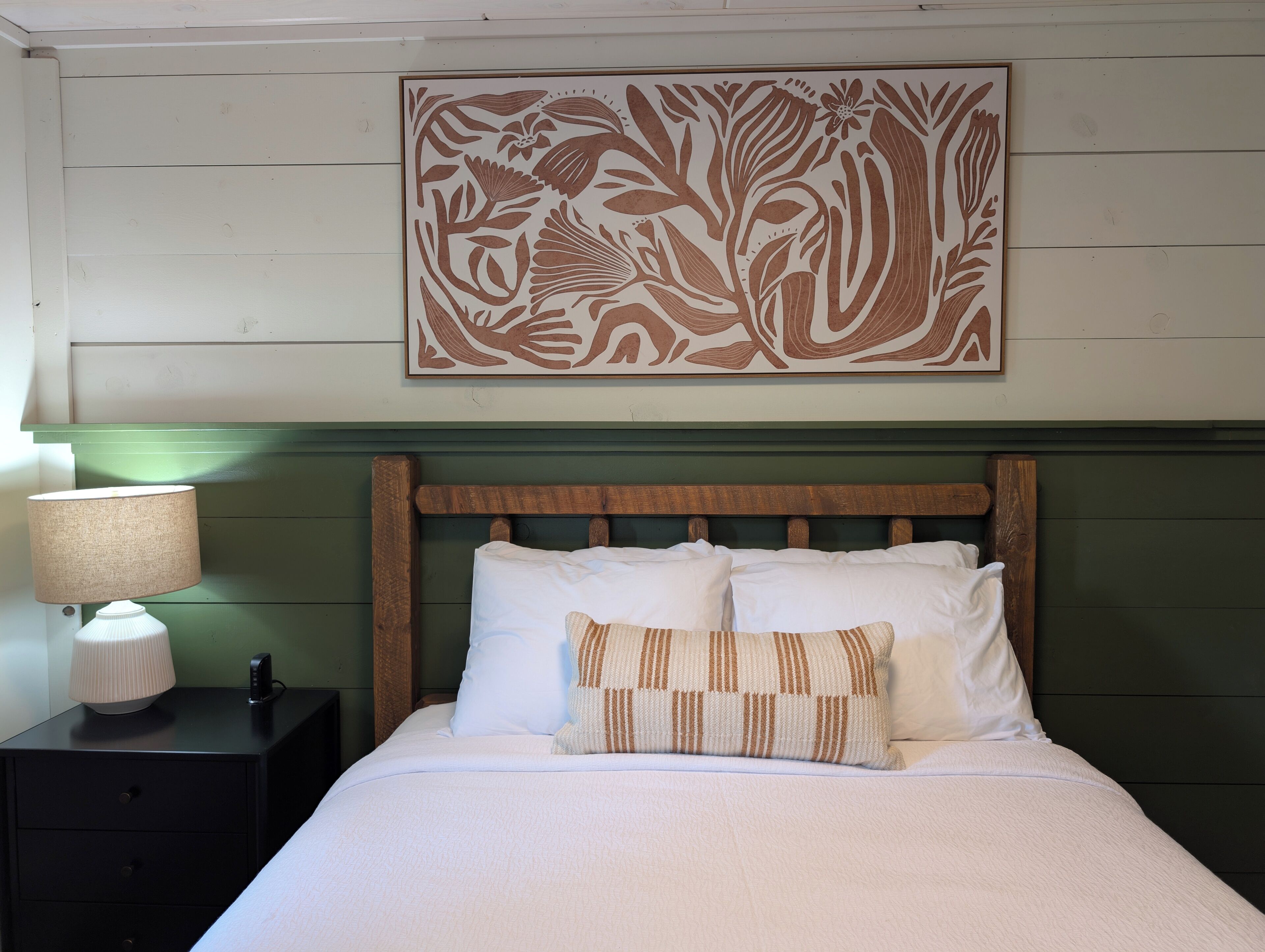 A neatly made bed with white bedding and a patterned pillow, a bedside lamp, and a wall-mounted artwork.