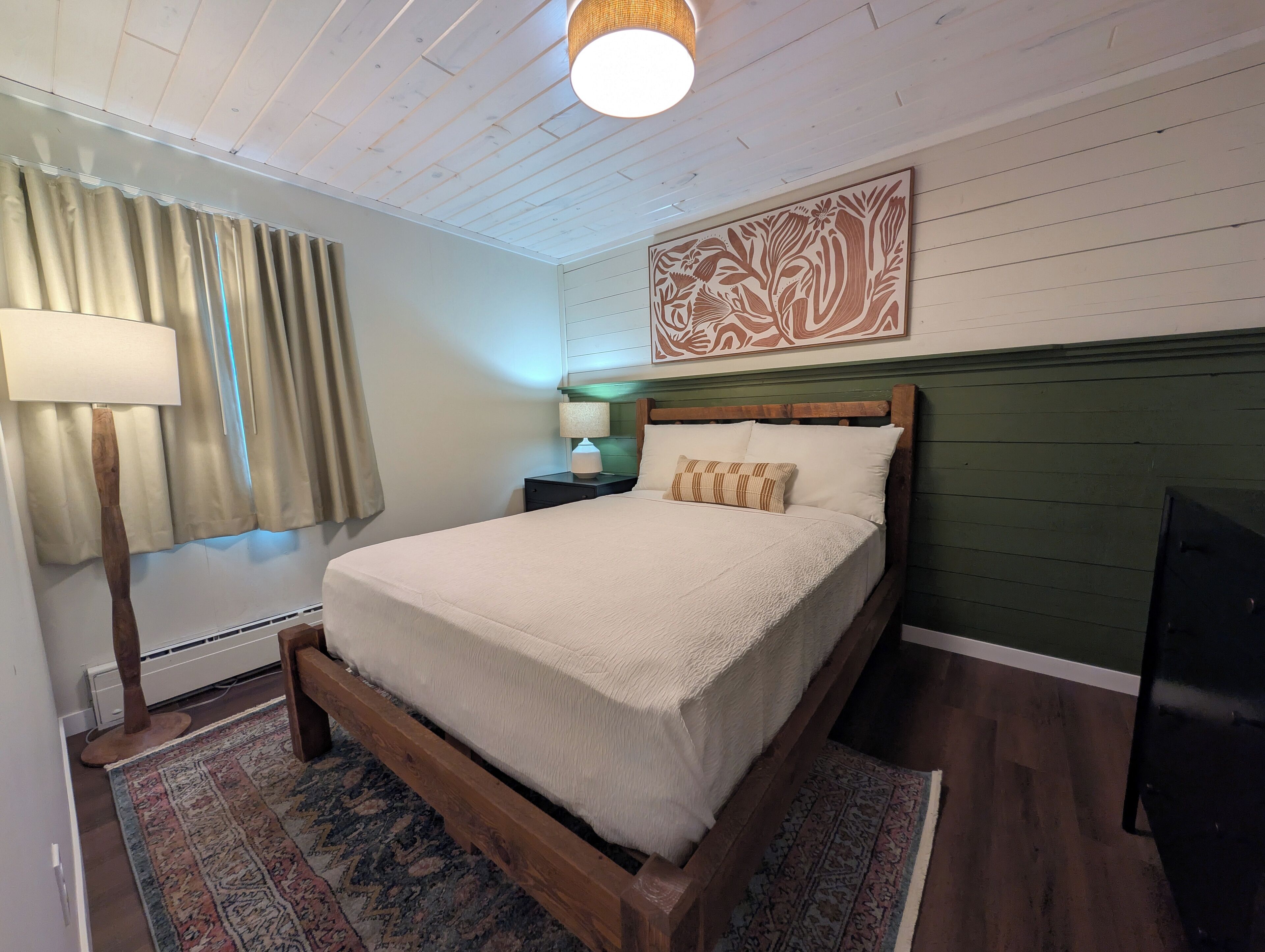A neatly arranged bedroom with a wooden bed frame, a patterned rug, a wall-mounted artwork, and a bedside lamp.