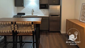 Une cuisine compacte équipée d’une cuisinière, d’un évier, d’un four à micro-ondes et d’un réfrigérateur. On y trouve une table avec deux chaises et un meuble contenant un tableau encadré.