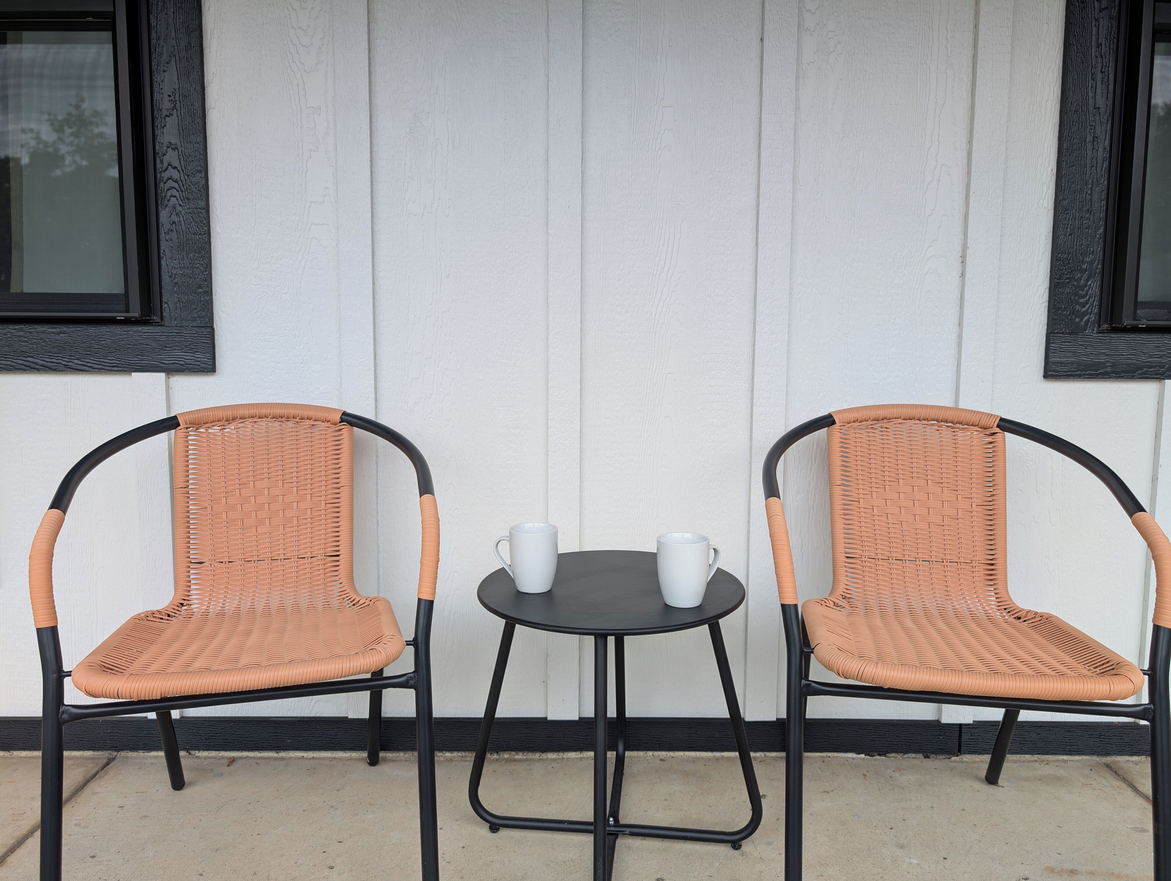 Two wicker chairs with black frames and a small round table with two white cups between them, against a white wall with black trim.