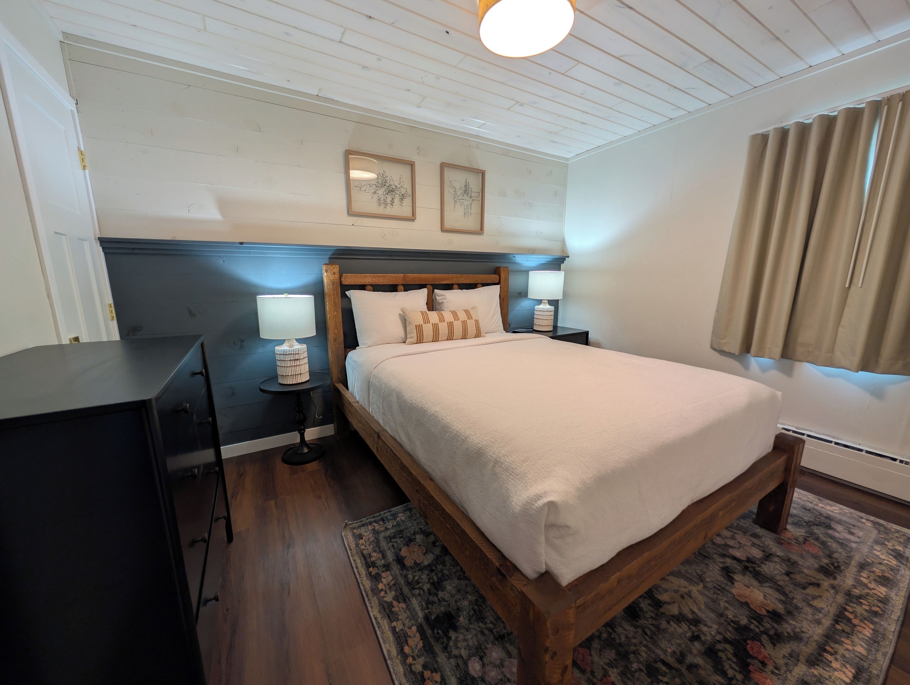 A bedroom with a wooden bed, two bedside lamps, a nightstand, a desk, and a patterned rug.