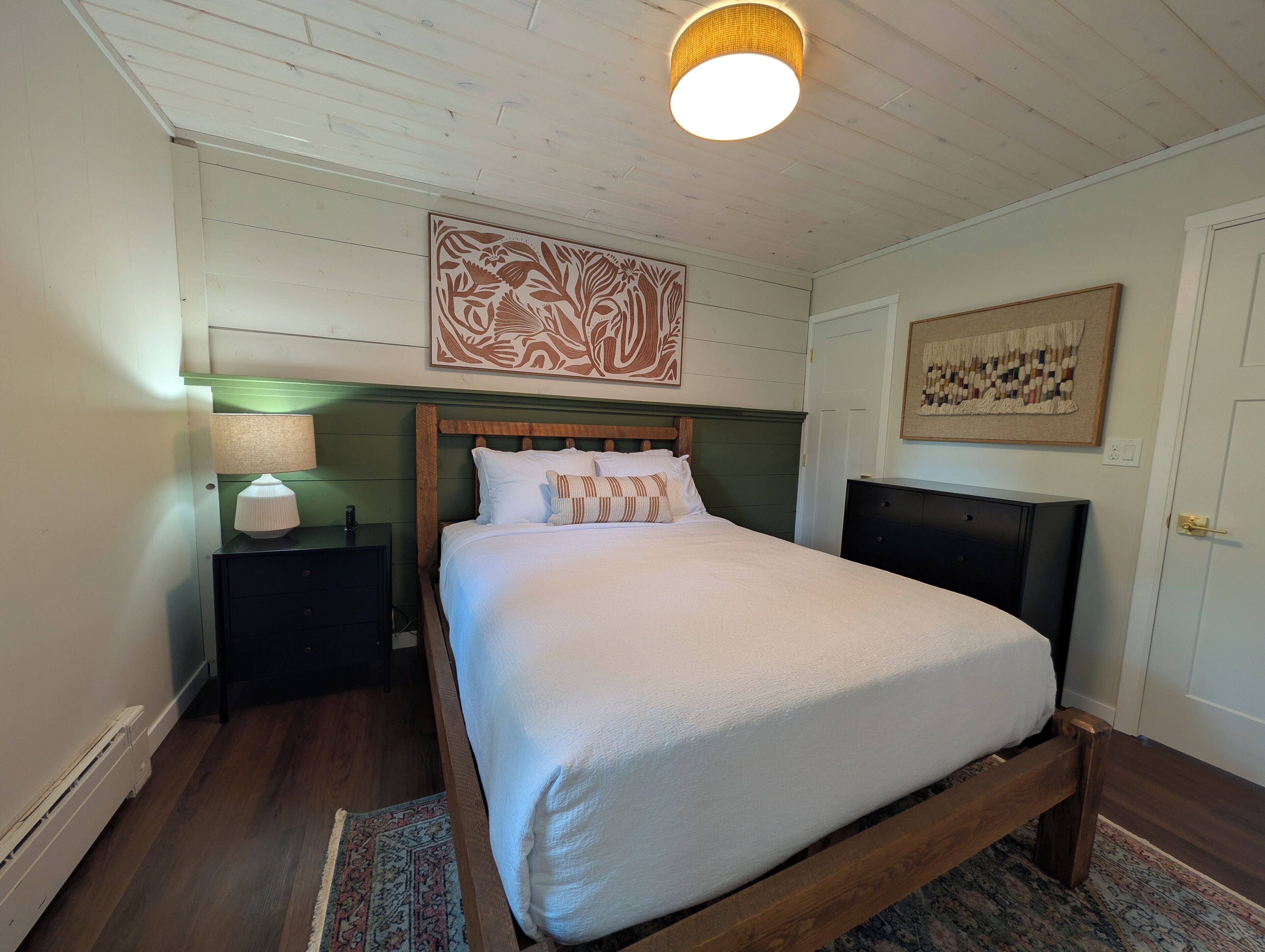 Een netjes ingerichte slaapkamer met een houten bed, een nachtkastje met een lamp, een decoratief muurkunstwerk en een kamerkleed met een patroon.
