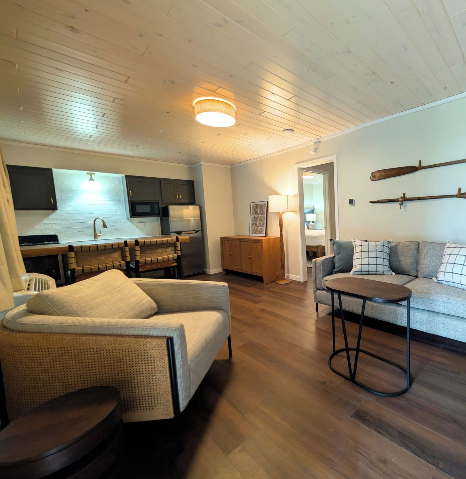 En moderne stue med sofa, lænestol og lille bord. Der er et køkkenområde med køleskab og skabe.