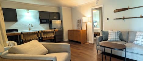 En moderne stue med sofa, lænestol og lille bord. Der er et køkkenområde med køleskab og skabe.