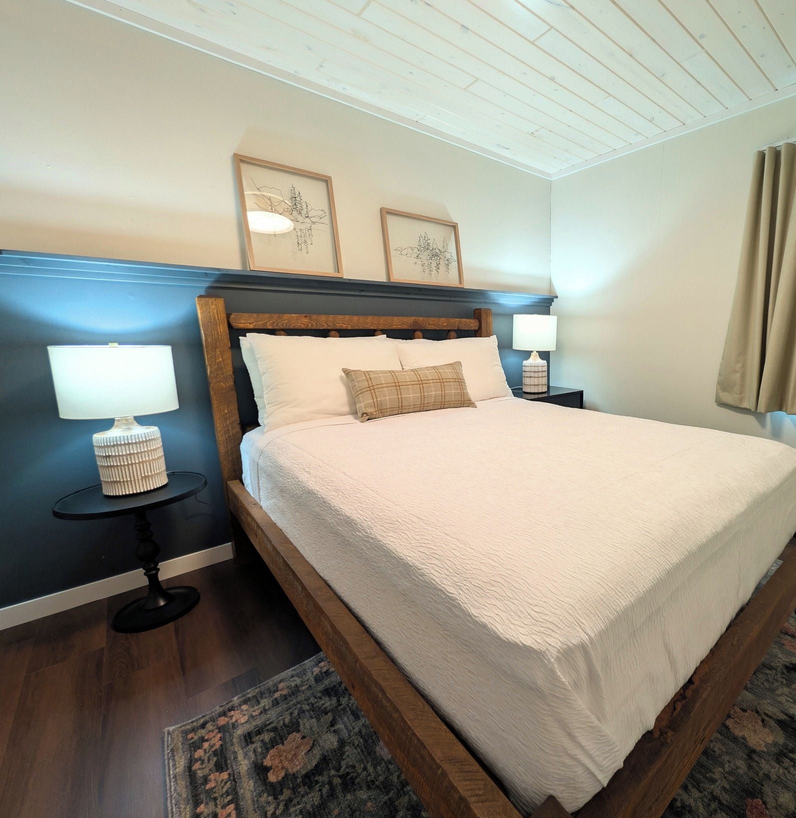 A neatly made bed with a white comforter and two pillows, flanked by two bedside tables with lamps, in a room with a wooden headboard and a large window with curtains.