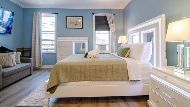 A bedroom with a bed, a dresser, a nightstand, a ceiling fan, and a window with curtains.