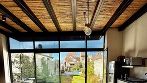 A modern kitchen with large windows, a dining area with a wooden table and chairs, and a view of a garden and buildings outside.