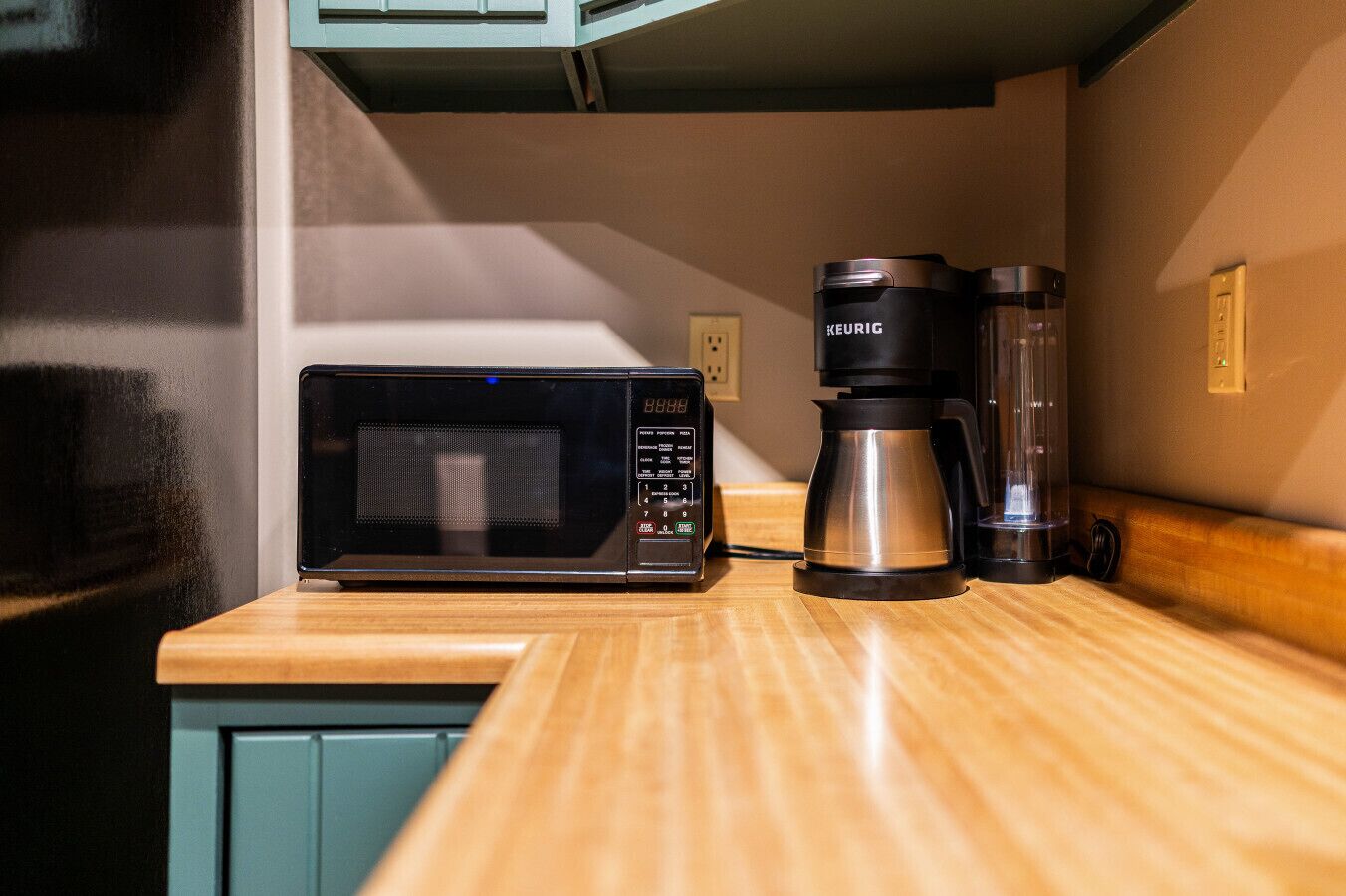 Una kitchenette con microondas, cafetera y un recipiente de vidrio sobre una encimera de madera.