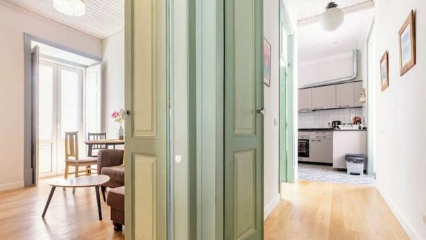 Un pasillo con una puerta verde, suelo de madera, un comedor con mesa y sillas, y una cocina al fondo.