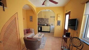 A compact living space with a sofa, armchair, coffee table, and a TV. A kitchen is visible through an archway. - Casa Flores. Historic center (Lagos)