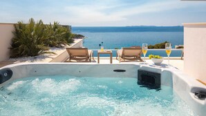 Ein privater Whirlpool mit zwei Liegestühlen und einem Tisch, mit Blick auf ein klares, blaues Meer, Schiffe und entfernte Küsten.