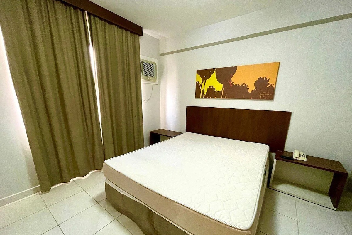 A hotel room with a bed, a nightstand, a telephone, a painting on the wall, and a window with curtains.