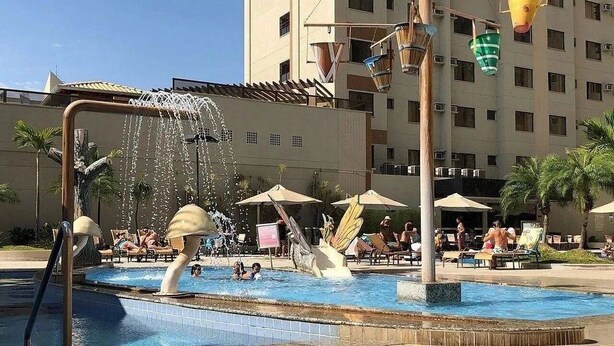 A swimming pool with a water fountain, lounge chairs, umbrellas, and a tall building in the background. - Boulevard Suíte Hotel (Caldas Novas)