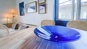 Un comedor con una mesa redonda que tiene un centro de mesa de vidrio azul, mobiliario de mimbre y arte náutico enmarcado en la pared.