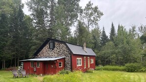 A red wooden cabin with a stone facade, surrounded by a grassy yard and dense forest. - Delightful cordwood cabin -Westmore near Burke Mtn, Lake Willoughby - Jay Peak. (Westmore)