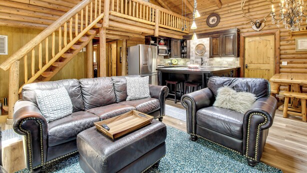 Ein Blockhaus-Interieur mit einer Holztreppe, Ledersofas, einer Küchenzeile und einer Deckenleuchte.
