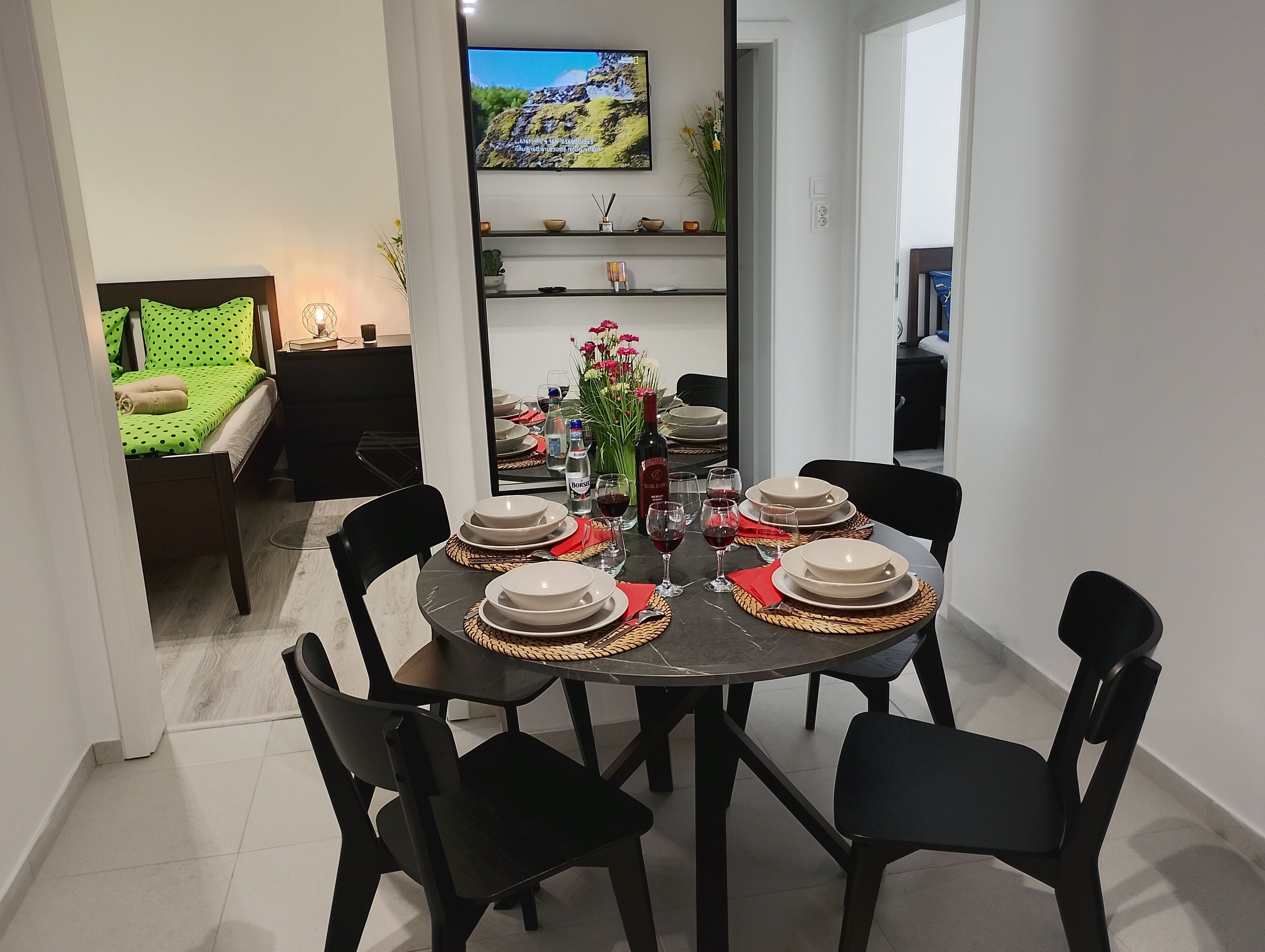 A dining area with a round table set for four, surrounded by black chairs. There is a TV mounted on the wall above the table, and a bed with green polka dot bedding is visible in the background.