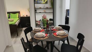 A dining area with a round table set for four, surrounded by black chairs. There is a TV mounted on the wall above the table, and a bed with green polka dot bedding is visible in the background.